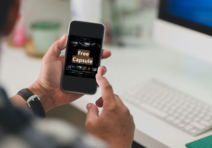 A person holding a smartphone with a website or app screen showing the text 'Free Capsule' in a blurred office setting with a keyboard and monitor in the background.
