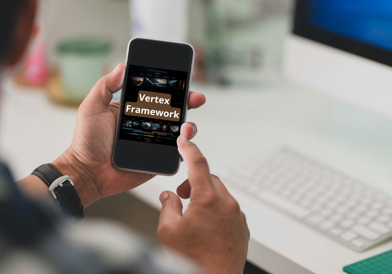 A person holding a smartphone showing the text 'Vertex Framework' on the screen, with a blurred desk and computer keyboard in the background.